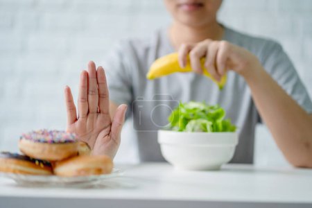 Téléchargez les photos : Jeune femme rejetant la malbouffe ou les aliments malsains tels que le beignet et choisissant des aliments sains tels que la pomme verte et la salade pour sa santé. - en image libre de droit