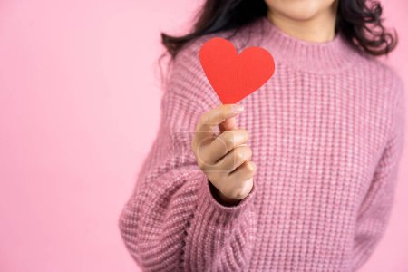 Téléchargez les photos : Closeup shot of female hand, beautiful asian woman wearing pink sweater is holding red love heart standing isolated on pink background, love concept - en image libre de droit