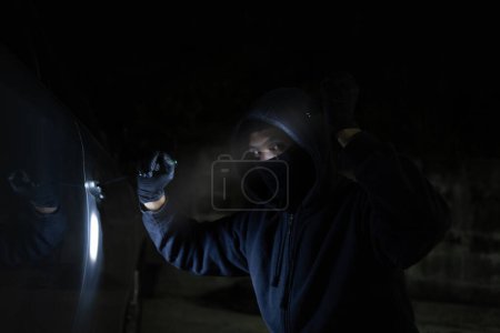 Photo for Young thief in black is stealing a car at night. He carries a flashlight and a screwdriver for car theft. - Royalty Free Image