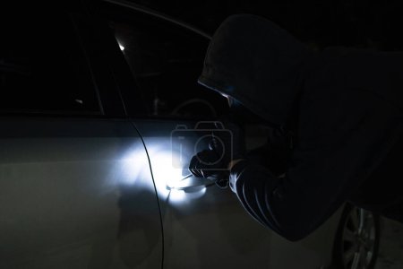 Photo for Asian man thief in black mask trying to open car using custom tool and flashlight. Car robber. - Royalty Free Image