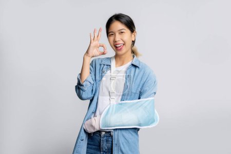 Foto de Hermosa trabajadora de oficina asiática con pantalones vaqueros azules se puso una férula suave debido a un brazo roto que muestra signo de bien en el fondo blanco, seguro de vida y accidente - Imagen libre de derechos