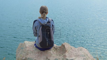 Photo for A woman tourist with a backpack on her shoulders sits on the top of a mountain and looks at the ocean. - Royalty Free Image