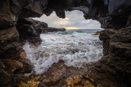 Téléchargez les photos : Une vue sur la mer orageuse à travers une vue à travers un rocher de lave au lever du soleil. Côte ouest de l'île de La Palma - en image libre de droit