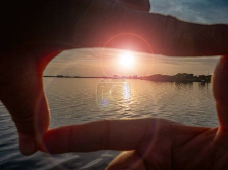 Foto de Gesto formado por dedos sobre el sol poniente en el mar. Lensflare desde el sol bajo sobre el mar tranquilo - Imagen libre de derechos