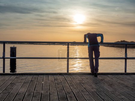 Foto de La turista se inclina sobre la barandilla y mira la puesta de sol sobre el mar. Muelle de mar o puente, noche Mar Báltico - Imagen libre de derechos