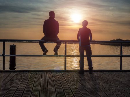 Foto de Un par de turistas. Una mujer se inclina sobre la barandilla y mira la puesta de sol sobre el mar. Un hombre se sienta en una barandilla y se relaja. Puente marítimo, Ruegen - Imagen libre de derechos