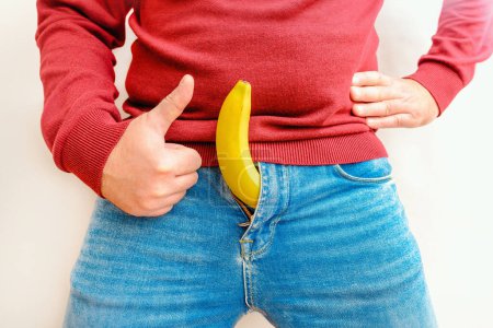 Foto de Hombre en pantalones vaqueros azules con plátano saliendo de sus pantalones, dando un pulgar hacia arriba contra fondo blanco.. - Imagen libre de derechos