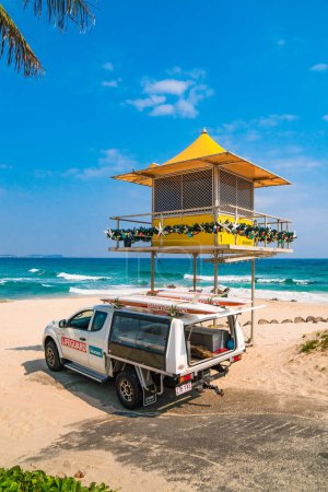 Foto de Camión salvavidas con tablas de surf en el techo aparcado junto a la torre de observación amarilla, y dos mujeres sentadas a la sombra de ella. Hermosa vista de la playa de Rainbow Bay en la Costa de Oro en Australia. - Imagen libre de derechos