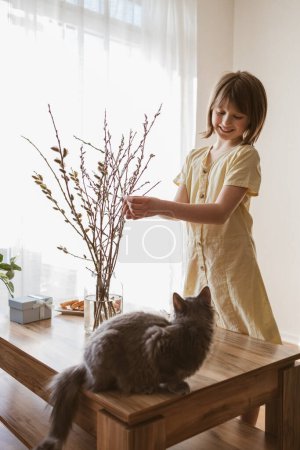 Foto de A happy girl hangs Easter eggs on a willow bouquet at home with a little kitten. Funny pets. Easter decoration. - Imagen libre de derechos