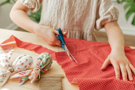 A girl at home makes Easter egg decorations in the shape of a rabbit from fabric. The child cuts the fabric with scissors. Ideas for home decoration.