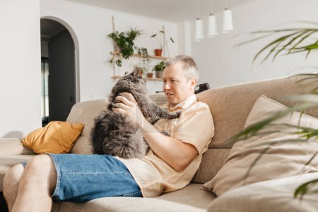 Photo for A middle-aged man sits and cuddles with a gray fluffy cat on the sofa in the living room and relaxes after work. Adored pets. - Royalty Free Image