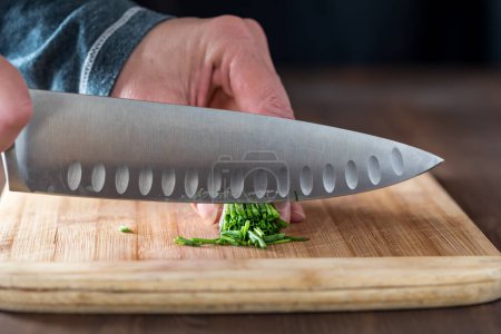 Photo for Close up of a chefs hands cutting a bundle of chives on a wooden board with a sharp knife. - Royalty Free Image