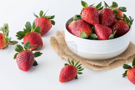 A bowl full of ripe juicy strawberries with several scattered about on the table.