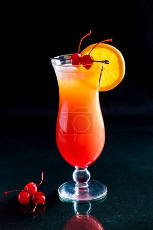 Tequila-Sonnenaufgang-Cocktail mit Orangenscheibe und Maraschino-Kirschen vor schwarzem Hintergrund.