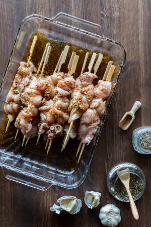 Photo for Above view of a glass dish containing raw chicken skewers in marinade on a dark wooden table. - Royalty Free Image