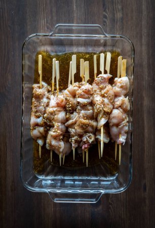 Photo for A glass dish of raw chicken marinading on bamboo skewers, on a dark wooden table. - Royalty Free Image