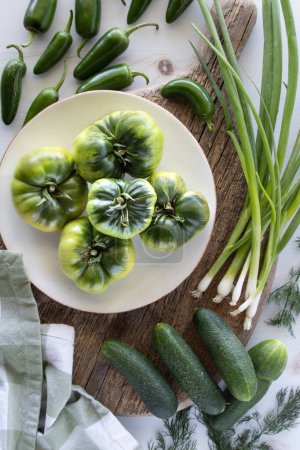 Foto de Tomates herederos verdes en un plato rodeado de verduras verdes frescas. - Imagen libre de derechos