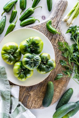 Foto de Vista de arriba hacia abajo de tomates herederos verdes en una tabla rústica, rodeado de otras verduras verdes. - Imagen libre de derechos