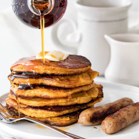 Photo for Syrup being poured onto a freshly made stack of sweet potato pancakes. Served with sausages. - Royalty Free Image