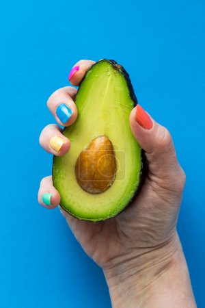 A hand with colourful nail polish holding a fresh cut avocado, against a bright blue background.