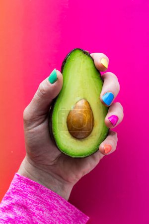 A hand with colourful nail polish holding a freshly cut avocado, against a bright pinky orange background.