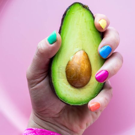 A hand with colourful nail polish holding half of a freshly cut avocado.