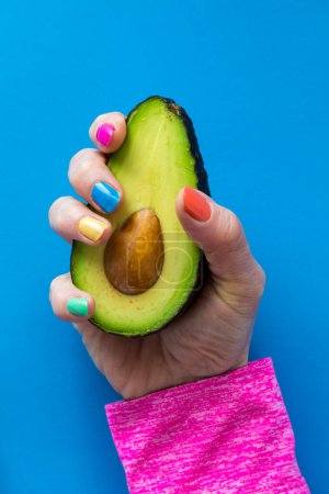 A hand with colourful nail polish holding a cut avocado, against a bright blue background. 