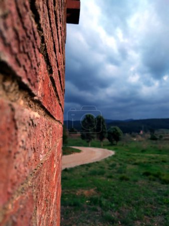 Foto de From a red brick wall illuminated by the light of day, a path with some trees can be seen in a landscape with a cloudy sky that threatens rain. - Imagen libre de derechos