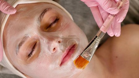 cosmetólogo profesional aplica una máscara blanca en la cara de una cliente femenina con un cepillo, primer plano