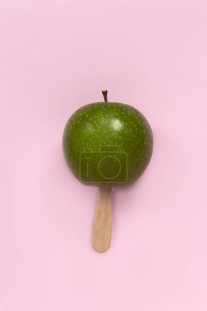 Téléchargez les photos : Pomme verte sur le bâton de crème glacée sur un fond rose pastel - en image libre de droit