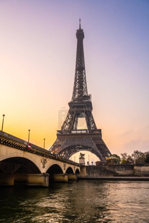 A captivating view of the Eiffel Tower and a bridge over the Seine River at sunrise. The sky is painted with warm hues, casting a serene atmosphere over Paris, France.