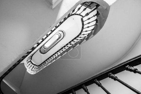Ein fesselndes Schwarz-Weiß-Bild, das das komplizierte Design einer Wendeltreppe einfängt und seine längliche ovale Form und sein detailliertes Geländer betont. Die Perspektive bietet einen faszinierenden Blick, dass
