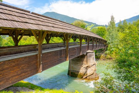 A long, covered wooden bridge spans a clear mountain stream surrounded by lush green foliage in a serene valley. Austria