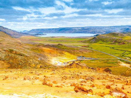 Ein lebhafter Sommerblick auf das geothermische Gebiet Seltun in der Nähe von Krysuvik mit farbenfrohem mineralreichen Gelände, heißen Quellen und einer Bergkulisse mit einem weit entfernten See unter einem teilweise bewölkten Himmel. Island