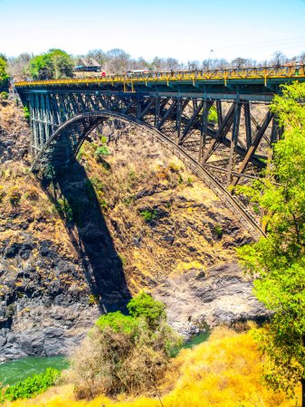 Una vista del icónico Puente de las Cataratas Victoria arqueándose sobre el abismo con exuberante follaje en el fondo y un cielo azul claro sobre la cabeza. Zimbabwe - frontera con Zambia. África