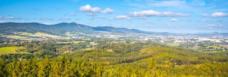 A sweeping vista of Liberec city nestled between rolling hills and Jested ridge under a blue sky dotted with clouds.