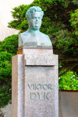A bronze bust of Viktor Dyk, prominently displayed on a stone pedestal, graces a lush park setting with green trees in the background. Vysehrad, Prague