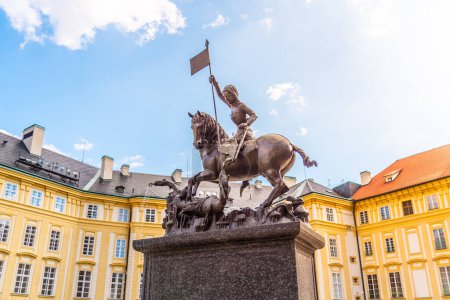 The bronze statue of Saint George triumphantly slays a dragon, set against the vibrant backdrop of Prague Castle under a blue sky. Prague, Czechia