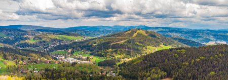 Ein Panoramablick auf den Tanvaldsky Spicak im Frühling, der den Hang, grüne Täler und eine kleine Stadt zwischen üppigen Wäldern unter wolkenverhangenem Himmel zeigt.