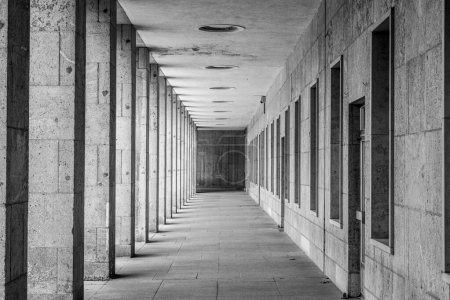 Ein verschwindender perspektivischer Blick auf eine moderne Arkade mit gleichmäßig angeordneten Säulen und Deckenleuchten. Schwarz-Weiß-Bild.