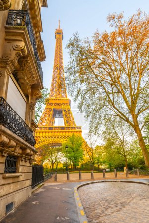 A breathtaking view of the Eiffel Tower illuminated during golden hour, surrounded by lush green trees and a classic Parisian building. The cobblestone path adds a touch of historic charm to the scene