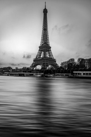 Ein fesselndes Schwarz-Weiß-Bild, das den ikonischen Eiffelturm vor einem wolkenverhangenen Himmel festhält, während die Seine im Vordergrund sanft fließt und die zeitlose Schönheit von Paris präsentiert