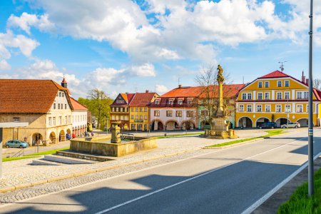 A picturesque view of a peaceful square in Zacler, Czech Republic. Colorful buildings with classic European architecture line the street. A beautiful fountain with an intricate statue is at the center