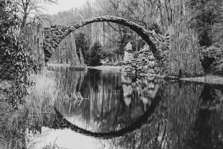 Rakotz Bridge, German: Rakotzbrucke, forms a perfect stone arch reflected in the calm waters of a serene German lake, surrounded by bare trees. Black and white image.