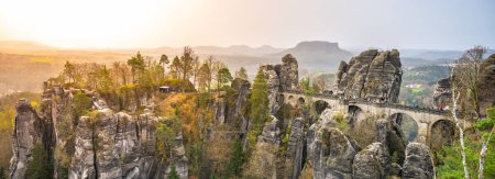Golden sunlight bathes the Bastei Bridge and surrounding sandstone rock formations at dawn in Saxon Switzerland, Germany.