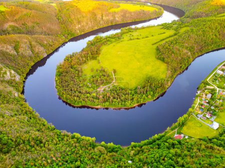 Una vista aérea captura la vibrante curva de herradura en el río Moldava en Solenice, Chequia. El río se curva dramáticamente a través de exuberantes paisajes verdes bajo un cielo despejado.