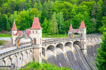 A view of the Les Kralovstvi Dam in Czechia, showcasing the intricate archway and stonework of the structure. The dam stands tall and imposing, surrounded by lush greenery.