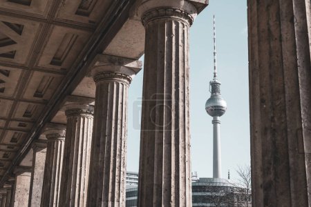 The Berlin TV Tower stands tall, framed by the classical columns of a nearby historical building on a clear day. Berlin, Germany