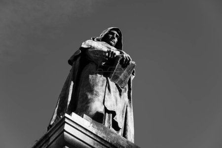 A black and white image of the Giordano Bruno Monument in Campo de Fiori, Rome, Italy. The statue is of a man in a robe, holding a book. It is popular tourist destination. Black and white photography.