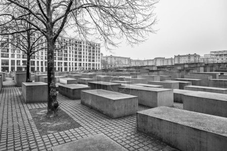Barren trees and concrete stelae stand under a cloudy sky at the Holocaust Memorial in Berlin. Black and white image.